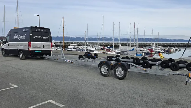 En stor bil med en påhenger står parked ved en marina med flere båter og båtplasser i bakgrunnen.